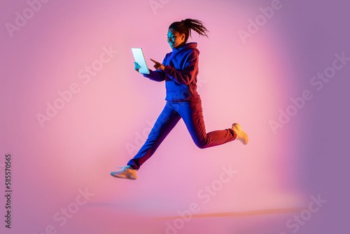 Full length of young black woman with tablet computer jumping in pink neon light background, copy space