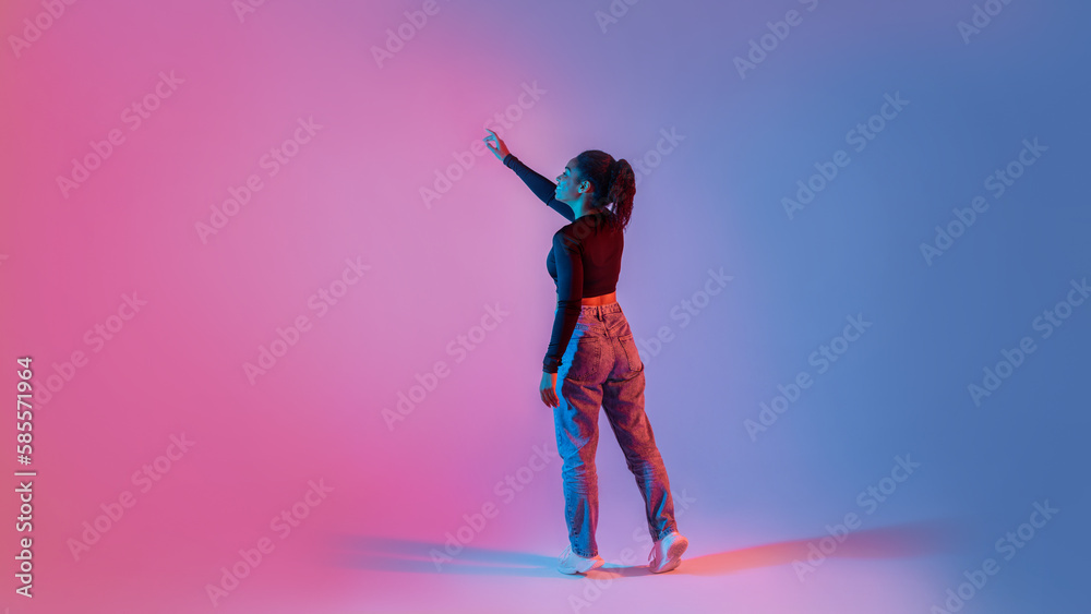 © Prostock-studio - Back view of young black lady using virtual interface, touching imaginary screen, standing on neon colorful background