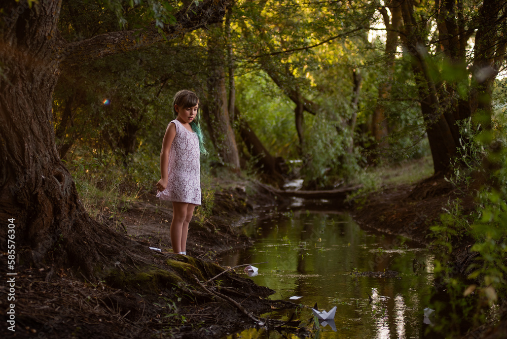 Teenage girl with green hair in dress, stands on the river bank, launches white paper origami boat Teenage girl with green hair in dress, stands on the river bank, launches white paper origami boat