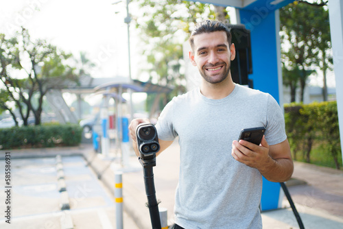 Cheerful happy Asian man using an EV charging application on smartphone to prepare vehicle charging and payment. Modern lifestyle of transportation with sustainability and sustainable energy. photo