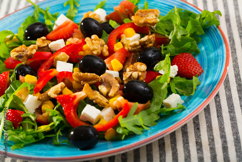 Fresh green arugula salad with vegetables, goat cheese and ripe strawberry decorated with black olives, walnuts and corn kernels