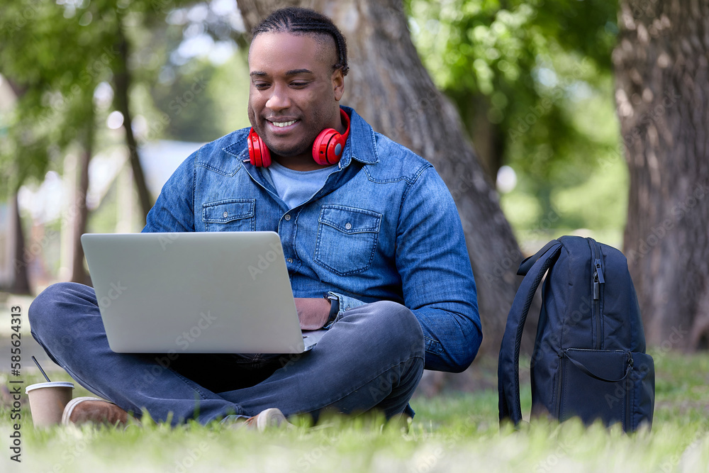 Stylish African businessman works on a laptop, analyzes statistical calculation data, sits on the grass and smiles. Digital entrepreneur is working online on a startup project Stylish African businessman works on a laptop, analyzes statistical calculation data, sits on the grass and smiles. Digital entrepreneur is working online on a startup project