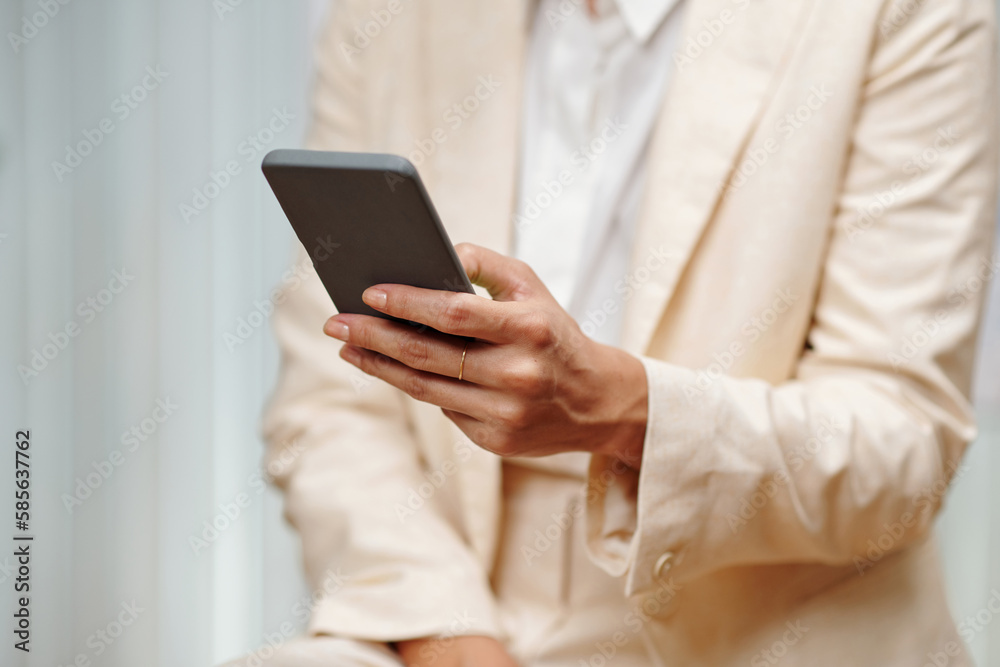 Hand of young woman in beige blazer texting in mobile phone or scrolling through contacts while standing in front of camera in office