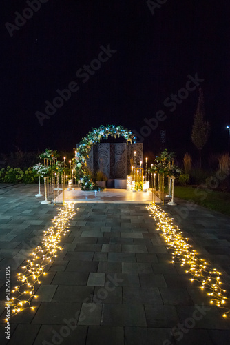 Stylish wedding round arch of candles, lanterns and fresh flowers, illuminated at night. Creative image for your design or illustrations.