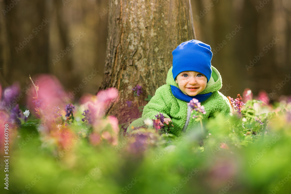 Baby boy among spring flowers in the forest. The child is dressed in a green overalls and a blue hat. Baby boy among spring flowers in the forest. The child is dressed in a green overalls and a blue hat.