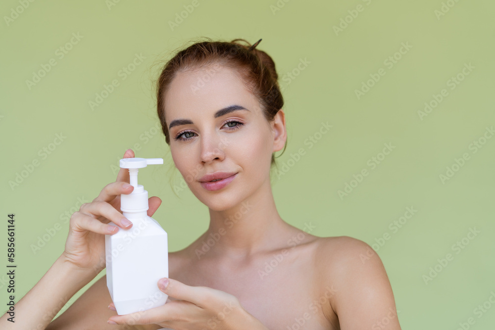 Beauty portrait of young topless red hair woman with bare shoulders on green background with perfect skin holding bottle of shampoo, body lotion Beauty portrait of young topless red hair woman with bare shoulders on green background with perfect skin holding bottle of shampoo, body lotion