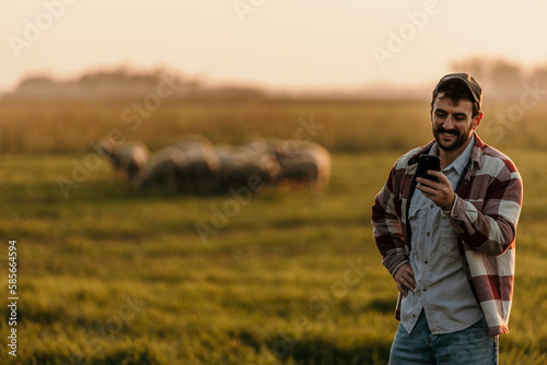 Male modern farmer using phone while watching a domestic animal herd on the horizon. Copy space © La Famiglia