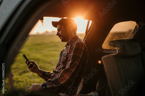 One man, a handsome gentleman sitting on a car trunk during the sunset, using a smartphone.