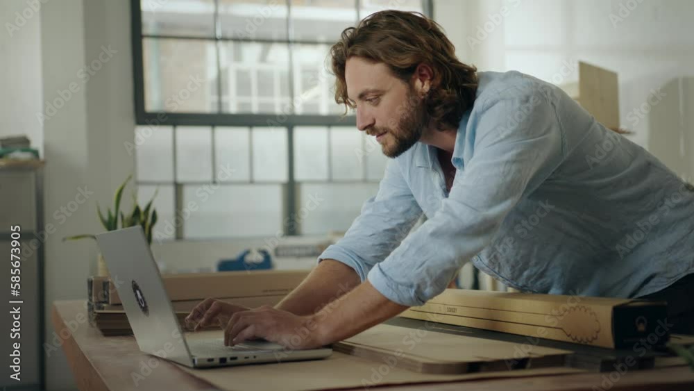 Focused young caucasian man in shirt leaning over merchandise while typing on laptop in office Focused young caucasian man in shirt leaning over merchandise while typing on laptop in office