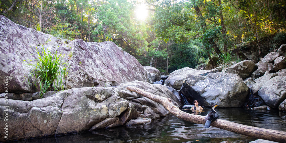 Fototapeta premium Beautiful girl takes a dip in cedar creek swimming holes and admires Australian pied cormorant sitting on a branch. Hidden gem near Brisbane, Queensland, Australia