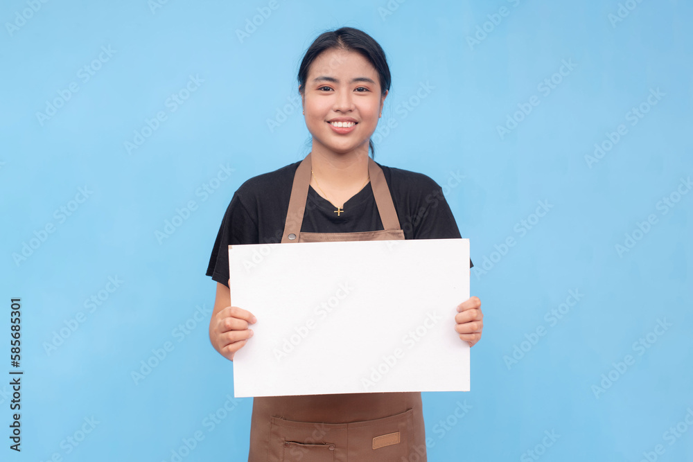 A young asian female waitress or barista holding a blank white signage. Wearing a brown apron and black shirt welcoming a customer and against a light blue background. A young asian female waitress or barista holding a blank white signage. Wearing a brown apron and black shirt welcoming a customer and against a light blue background.