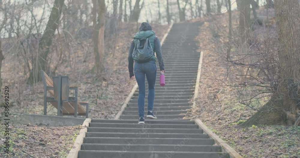 Weary young unrecognizable woman walks up concrete staircase with flask in her hand. City park, daytime, springtime, back view.