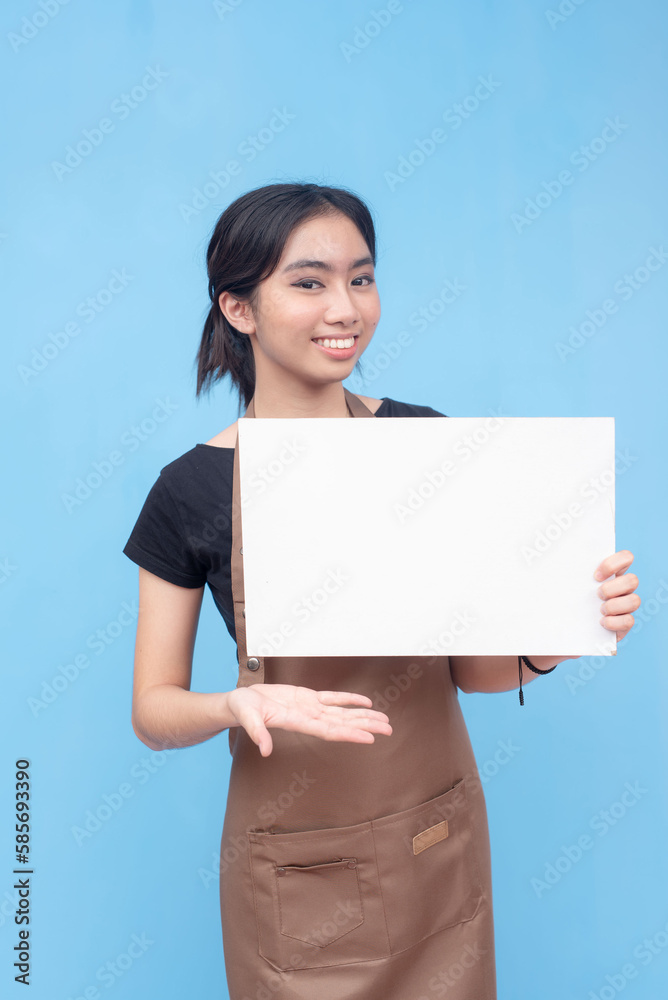A young asian female waitress or barista presenting a blank white signage. Wearing a brown apron and black shirt welcoming a customer and against a light blue background. A young asian female waitress or barista presenting a blank white signage. Wearing a brown apron and black shirt welcoming a customer and against a light blue background.