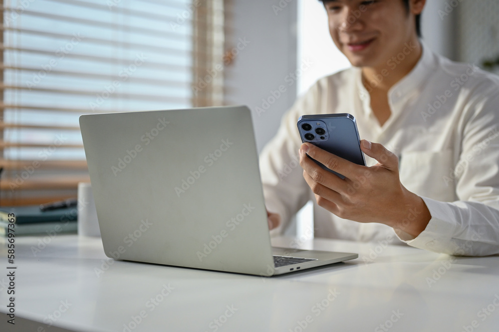 Cropped image of a handsome asian businessman using his phone and laptop at his desk.