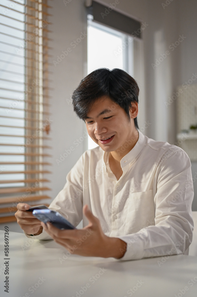Smiling millennial Asian man using mobile banking application to pay online bills.