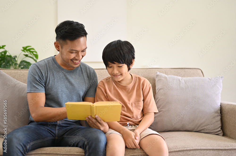Happy and kind Asian dad is telling a story to his son on a sofa in the living room Happy and kind Asian dad is telling a story to his son on a sofa in the living room
