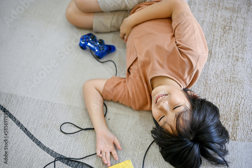 A happy cute Asian little boy laying on the living room floor with video game joysticks.