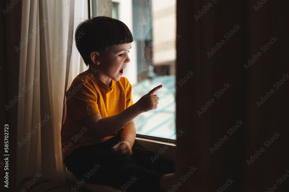 Little cute attractive boy sitting on windowsill and look at window, boy point his finger on something, what he saw on window. Kid look shocked amazement and surprised, he wear yellow t-shirt at home. Little cute attractive boy sitting on windowsill and look at window, boy point his finger on something, what he saw on window. Kid look shocked amazement and surprised, he wear yellow t-shirt at home.