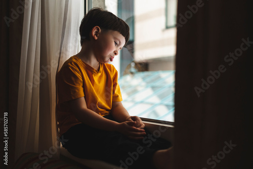 Fotografija Portrait of sad and gloomy little boy sulking from disappointment, sitting on windowsill upset and look at window