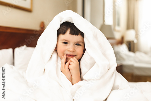 Little cute boy wrapped himself in a white blanket while sitting on the bed and smiling. Happy kid look surprised, his hands on chin, amazement toddler boy. Good morning.