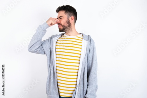 Young caucasian mán wearing trendy clothes over white background smelling something stinky and disgusting, intolerable smell, holding breath with fingers on nose. Bad smell