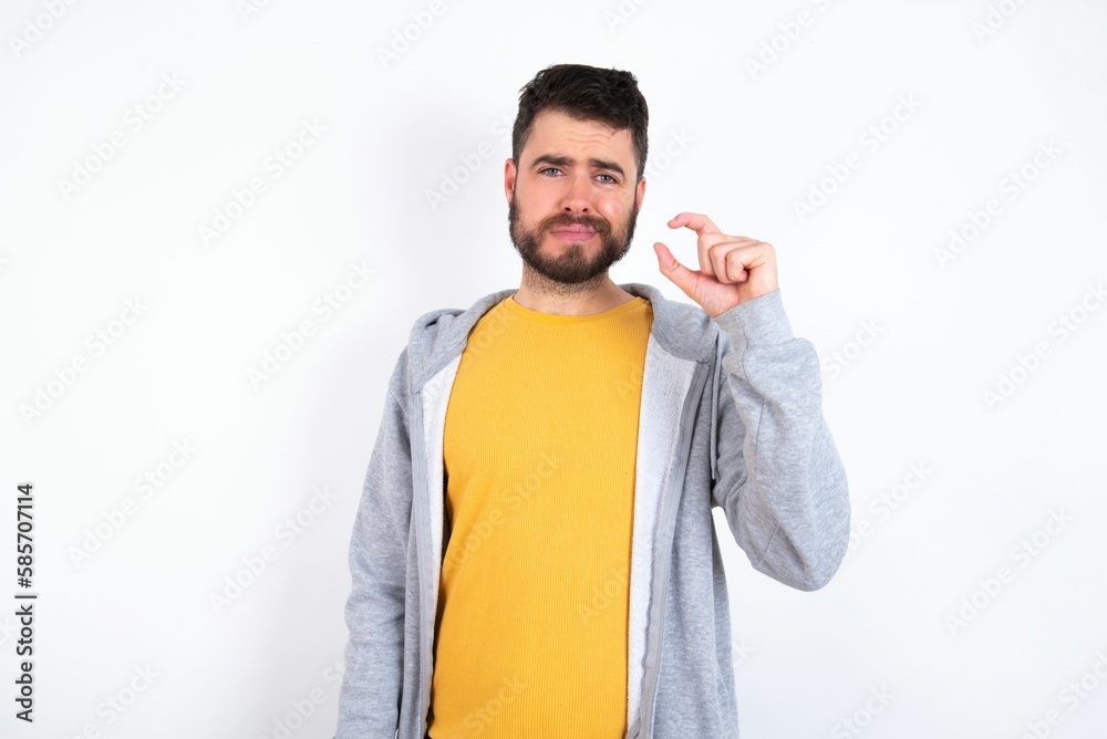 Displeased Young caucasian mán wearing trendy clothes over white background shapes little hand sign demonstrates something not very big. Body language concept. Displeased Young caucasian mán wearing trendy clothes over white background shapes little hand sign demonstrates something not very big. Body language concept.