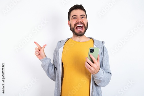 Astonished Young caucasian mán wearing trendy clothes over white background holding her telephone and pointing with finger aside at empty copy space