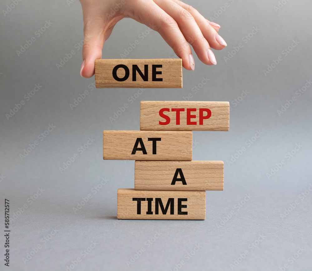 One step at a time symbol. Concept words One step at a time on wooden blocks. Beautiful grey background. Businessman hand. Business and One step at a time concept. Copy space. One step at a time symbol. Concept words One step at a time on wooden blocks. Beautiful grey background. Businessman hand. Business and One step at a time concept. Copy space.