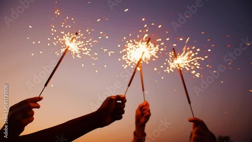 Group of people standing outdoors in the sunset with hands raised in the air, in which they holding lit sparklers with the sparks flying to the wind close up