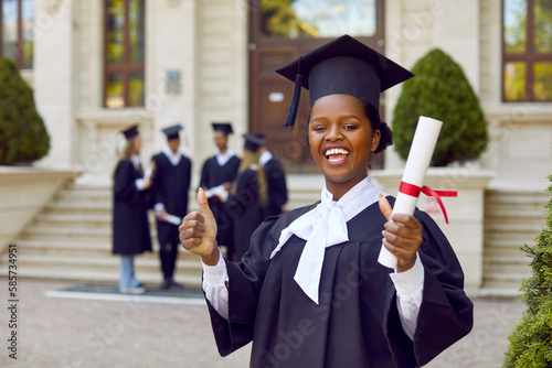 Portrait of happy cheerful joyful good successful beautiful African student girl in graduation cap and gown standing outside university building, holding diploma scroll, giving thumbs up and smiling