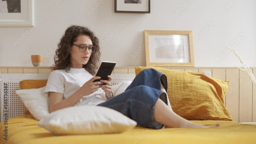 Pretty girl is reading a book in bright room at home. Beauty young woman enjoys reading and free time. Female student in glasses reads electronic book ebook in cozy bright living room, sitting on bed Pretty girl is reading a book in bright room at home. Beauty young woman enjoys reading and free time. Female student in glasses reads electronic book ebook in cozy bright living room, sitting on bed