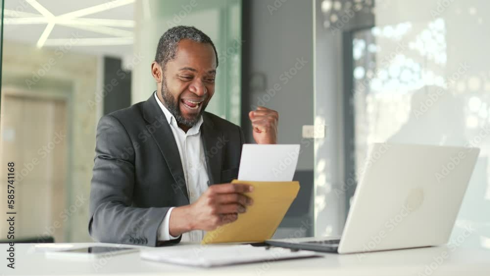 Happy african american businessman in formal suit reading letter with good news while sitting in office. Smiling male celebrates victory by holding hand in triumph gesture after receiving notification Happy african american businessman in formal suit reading letter with good news while sitting in office. Smiling male celebrates victory by holding hand in triumph gesture after receiving notification