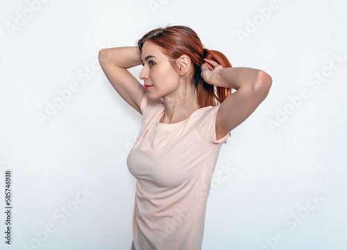 Young attractive woman fixing hair while holding hands behind head. Light T-shirt. White background.