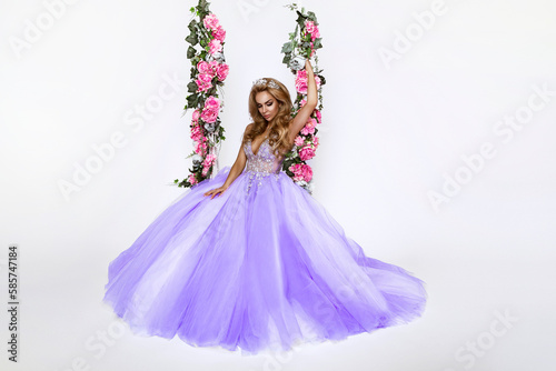 Beautiful natural woman in elegant long dress on a floral swing on white background. Glamour female model. Spring concept. Bridal fashion.