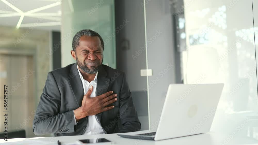 Mature african american man in formal suit suffering from tension and chest pain while sitting at desk at workplace in office. A sick male is having a heart attack. He needs urgent medical attention Mature african american man in formal suit suffering from tension and chest pain while sitting at desk at workplace in office. A sick male is having a heart attack. He needs urgent medical attention