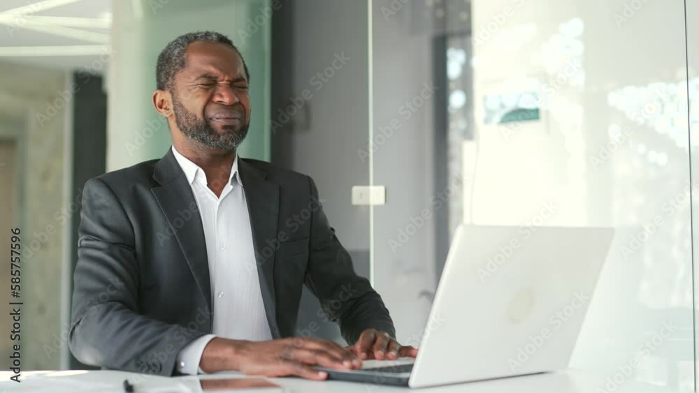 Mature african american businessman in formal suit suffering from back muscle pain while sitting at desk at workplace in modern office. Tired middle aged man is massaging his lower back with his hands Mature african american businessman in formal suit suffering from back muscle pain while sitting at desk at workplace in modern office. Tired middle aged man is massaging his lower back with his hands