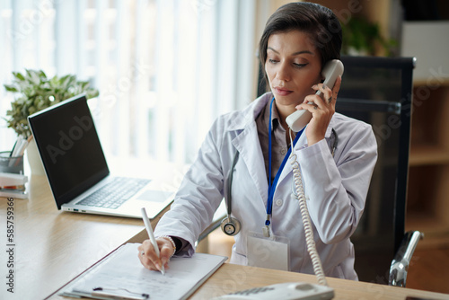 Fotografija Serious physician talking on phone with patient and writing down his complaint