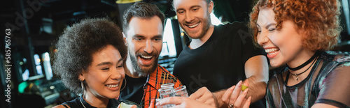 Fotografija Cheerful interracial friends holding tequila shots with salt in bar, banner