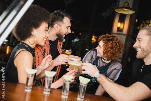 Side view of multiethnic friends with different cocktails talking near blurred tequila glasses in bar.