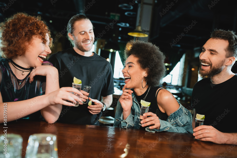 Carefree interracial people holding tequila shots near stand in bar.