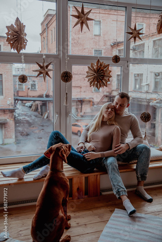 Beautiful young couple in love at the window hugging, next to their dog