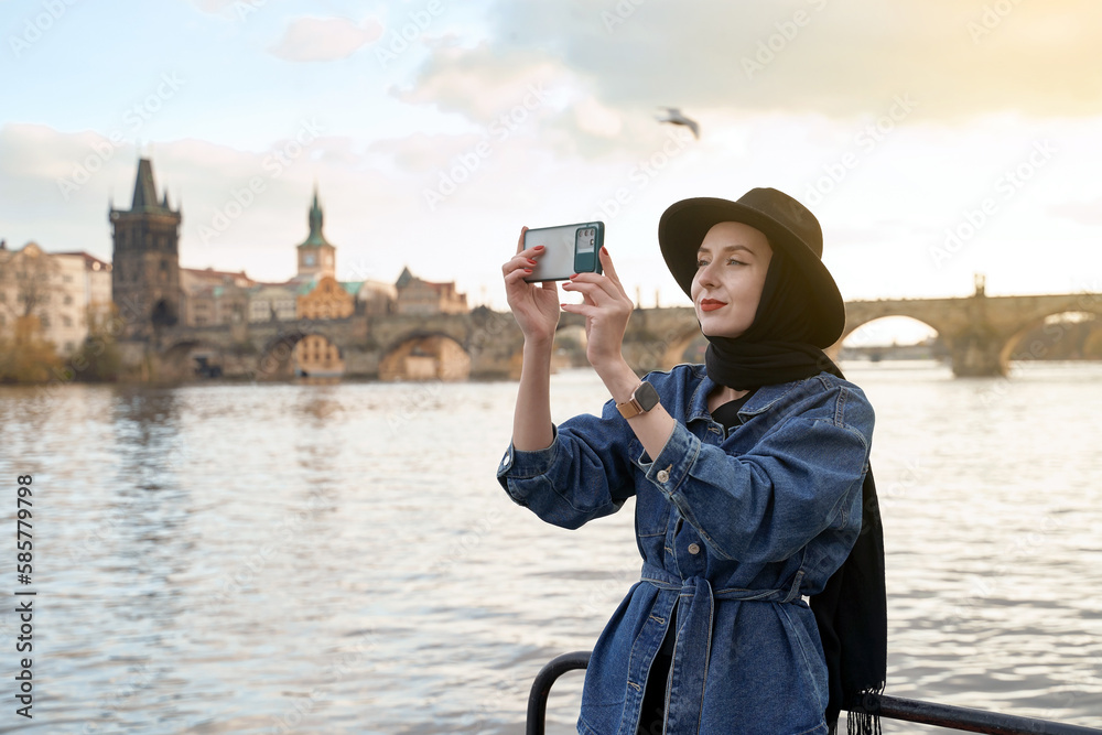 Stylish young woman traveller wearing black hat sitting taking pictures with her smartphone on Vltava river shore in Prague with Charles Bridge on background.
