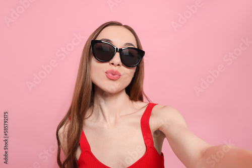 Beautiful young woman with sunglasses blowing kiss while taking selfie on pink background