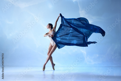 Beautiful ballerina wearing beige bodysuits emotional dancing with fabric silk over blue background