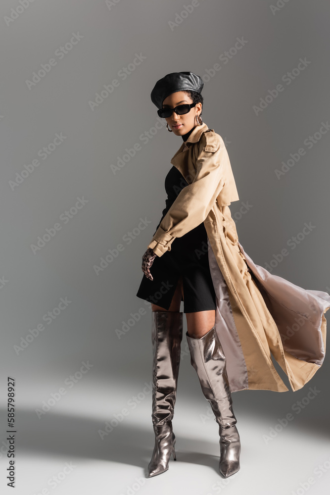 Full length of fashionable african american woman in sunglasses and trench coat on grey background. Full length of fashionable african american woman in sunglasses and trench coat on grey background.