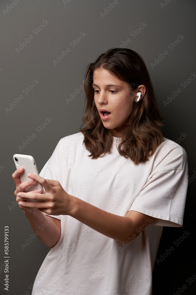 Surprised teen female in earbuds and looking at smartphone in hands, standing on dark background Surprised teen female in earbuds and looking at smartphone in hands, standing on dark background