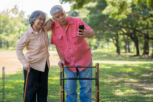 An old elderly Asian man uses walker and use smartphone for taking photos with his wife. Concept of happy retirement With Love and care from family and caregiver, Savings, and senior health insurance