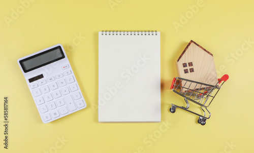 flat layout of wooden house model in shopping trolley, white calculator, blank page opened notebook on yellow background with copy space, home purchase concept.