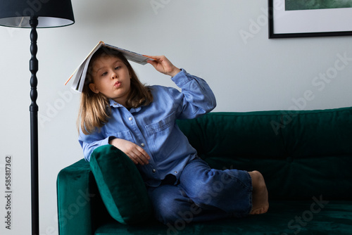 portrait of a schoolgirl in blue shirt sitting on sofa with book on head, funny face
