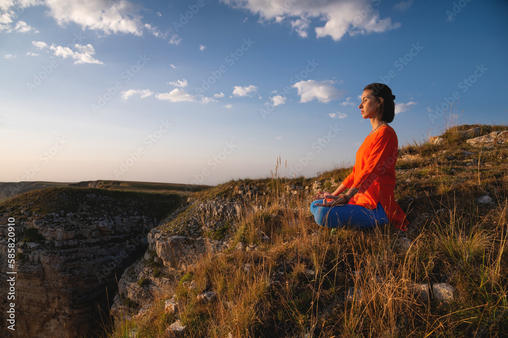 Panoramic view. young woman sitting on a grassy hill in lotus position while meditating outdoors in the morning. Sports woman doing meditation yoga in the mountains at sunset